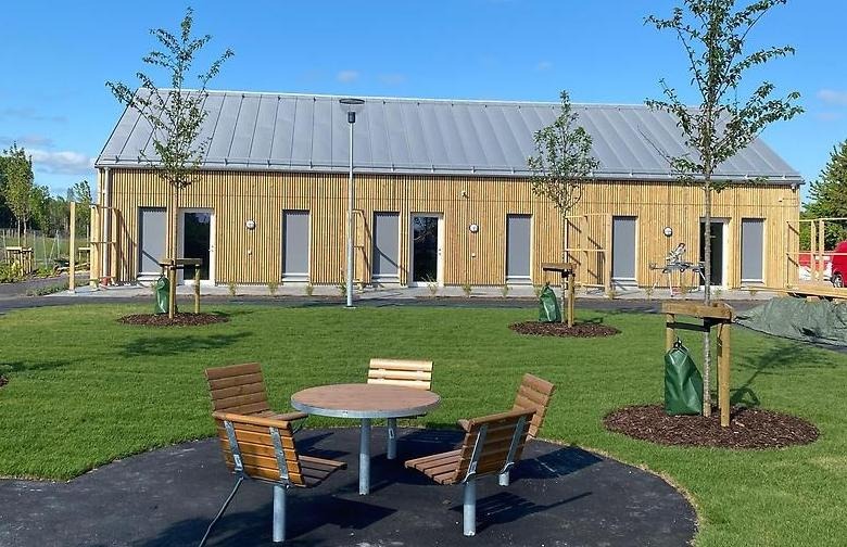 Fotografi på Lövgatans LSS-boende utemiljö. Ett runt träbord med fyra stolar i förgrunden, grön gräsmatta med nyplanterade träd och huset längst bak i bilden.