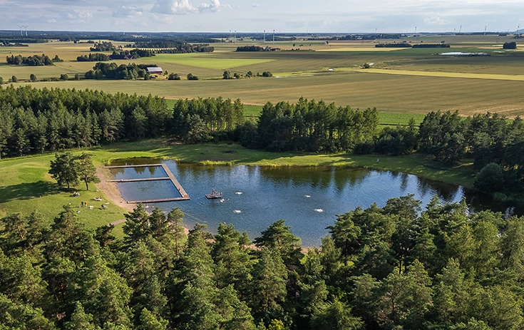 Drönarbild över en liten badsjö med bryggor och strand, omgiven av gräs, skog och åkermark.