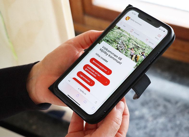 Händer som håller i mobiltelefon som visar Mjölby kommuns webbplats.