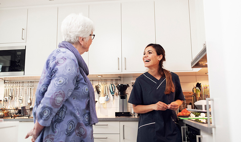 En äldre kvinna tillsammans med en yngre kvinna som står i ett kök. Hon har blå arbetsdräkt på sig. Båda tittar på varandra och ler.