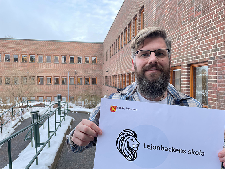Projektledare Kristoffer står framför stadshuset och håller upp en lapp med "Lejonbackens skola" skrivet på.