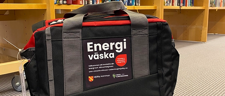 Svart väska med texten "Energiväska" på golv framför en bokhylla.