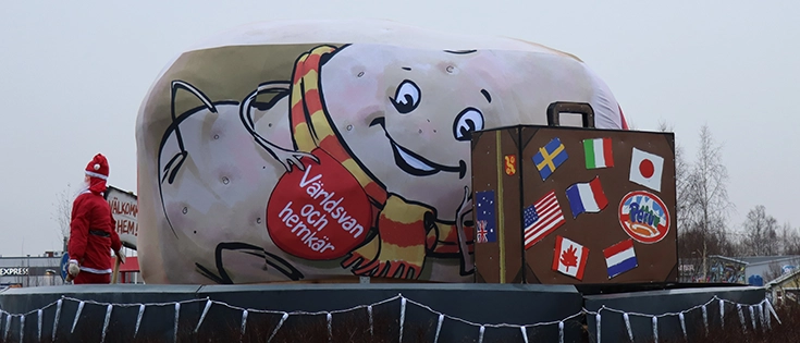 Stor konstpotatis draperad i tyg med illustration föreställande liggande potatiskaraktären Eddi. Framför en stor resväska med klistermärken på länders olika flaggor.