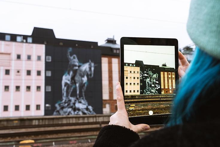 Man ser konstverk med AR-teknik, byggnaden med konstverket i bakgrunden. Person håller en läsplatta med appen nedladdad där bilden får liv. 