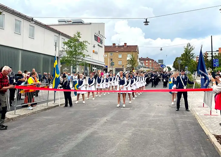 Ett rött band som hänger över en gata. Bakom bandet står en musikkår. Svenska flaggan.