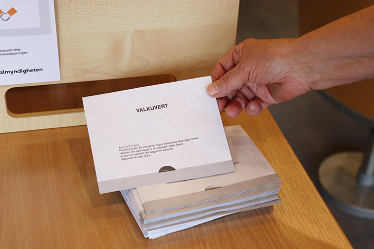 Hand håller upp valkuvert framför andra kuvert som ligger på ett bord.