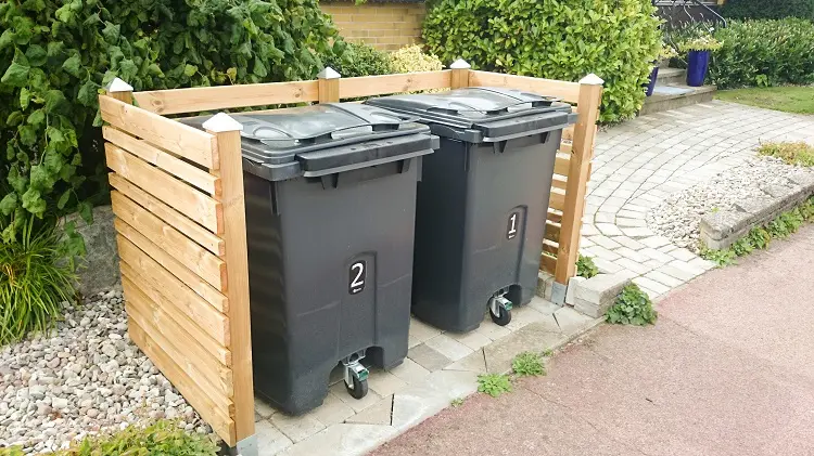 Två soptunnor med nummer 1 och 2 står i en trädgård, omgivna av ett U-format staket.