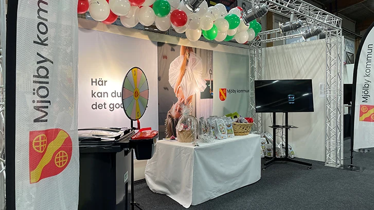 Upplyst mässmonter för Mjölby kommun med soptunnor, spelhjul, bord med korg och potatissäckar och en tv skärm.