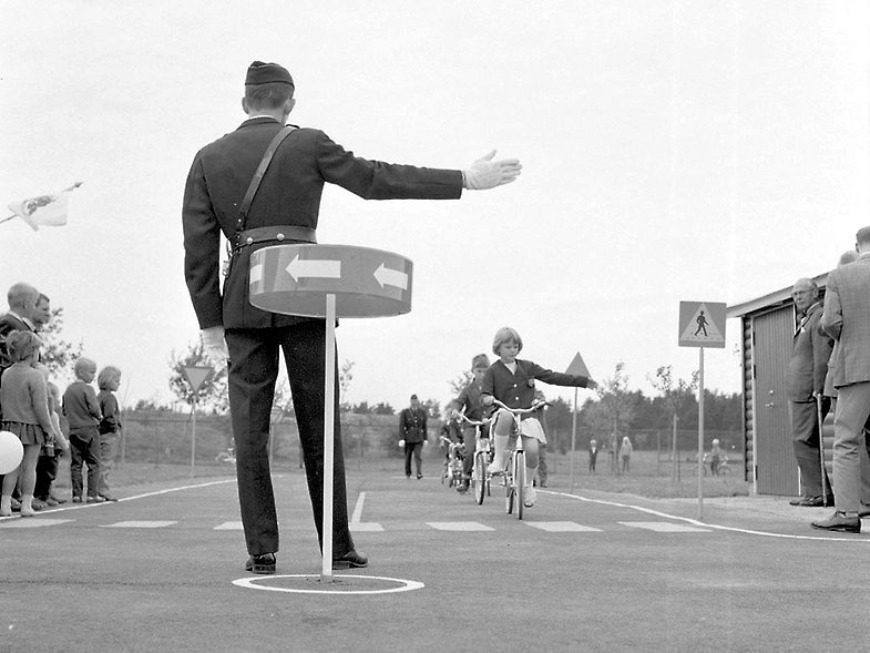 Svartvitt fotografi. Polis håller ut högerhanden framför cyklande barn på äldre lekplats.