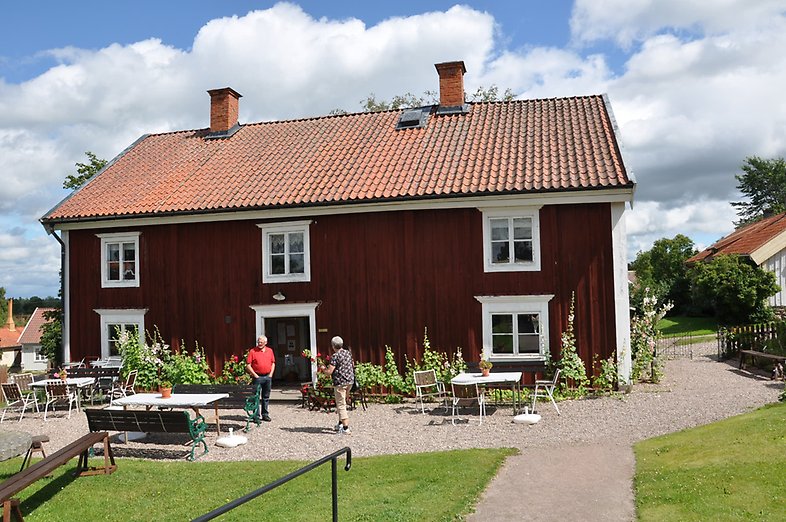 En röd stuga i två våningar med vita knutar. Två personer bland utställda bord och stolar.
