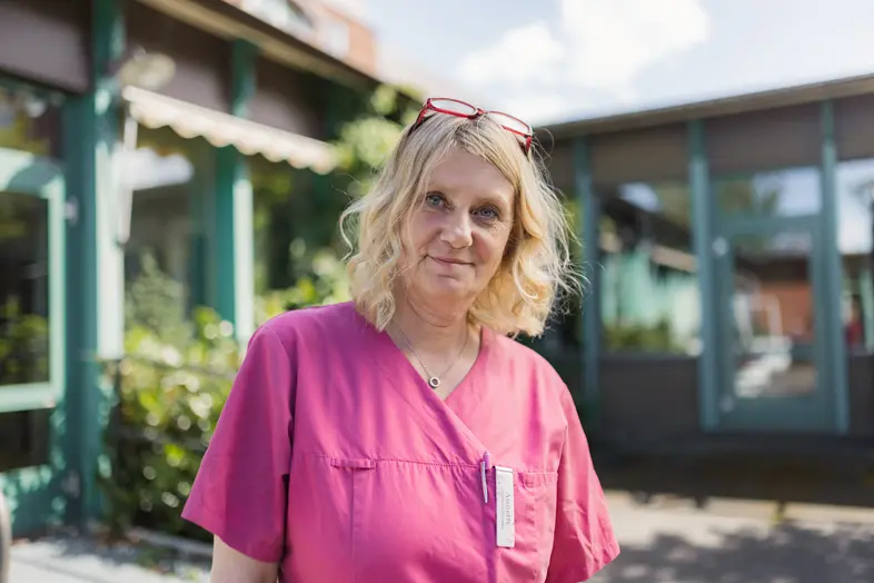 Kvinna i rosa arbetskläder framför en turkos byggnad i solljus.