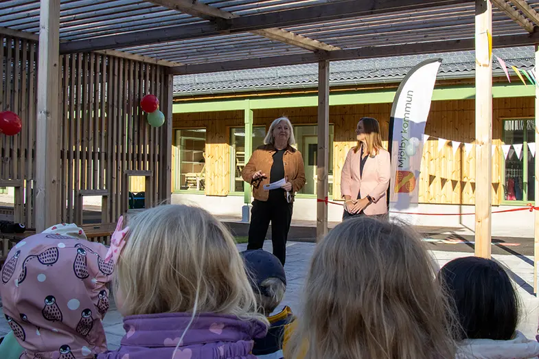 Två personer håller tal framför publik med barn.