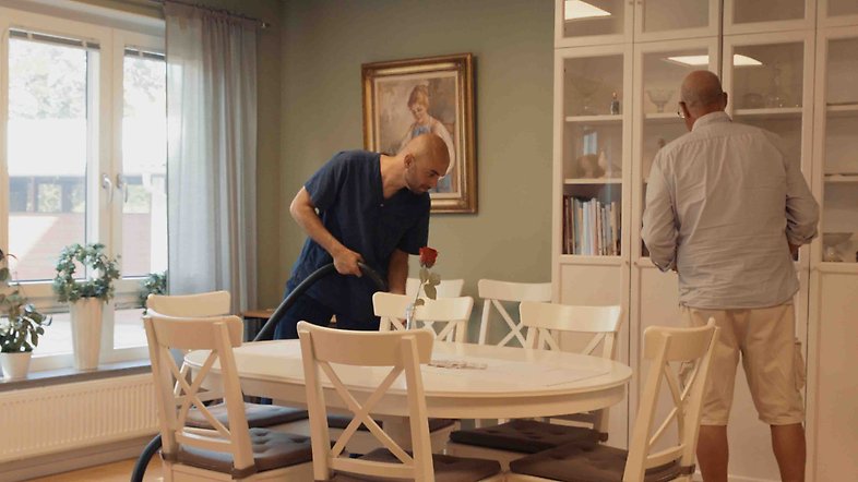 En äldre man städar sin lägenhet med en yngre manlig sommarvikarie.