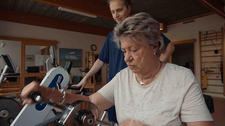 En äldre kvinna på ett särskilt boende tränar handcykel med en manlig, yngre medarbetare som stöd i bakgrunden.