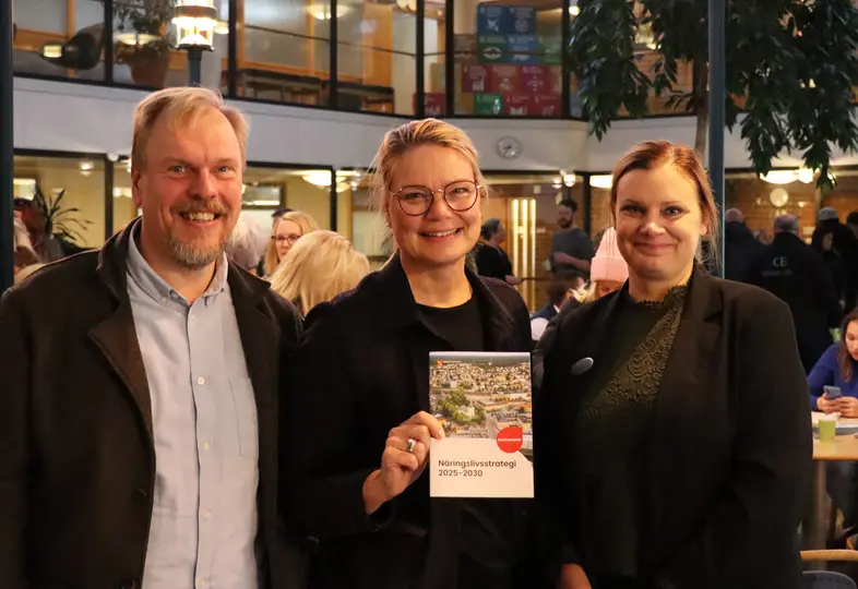 Tre personer tittar in i kameran, personen i mitten håller upp en broschyr.