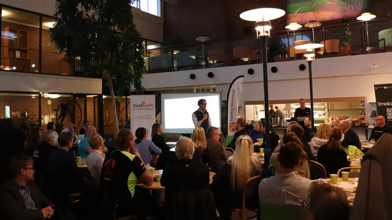 En person talar i mikrofon framför en publik. Publiken sitter samlade kring flera runda bord och tittar mot talaren.