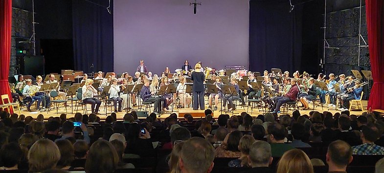 Ett 50-tal blåsarmusiker och en dirigent på en scen vid en konsert. Det är många i publiken och en del filmar med mobilen.