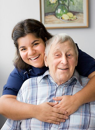 Kvinnligt vårdbiträde kramar om en äldre farbror, båda ler