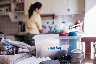 Låda med matvaror märkt "krislåda", vantar, ljus. En  person står och fyller på vatten i bakgrunden.